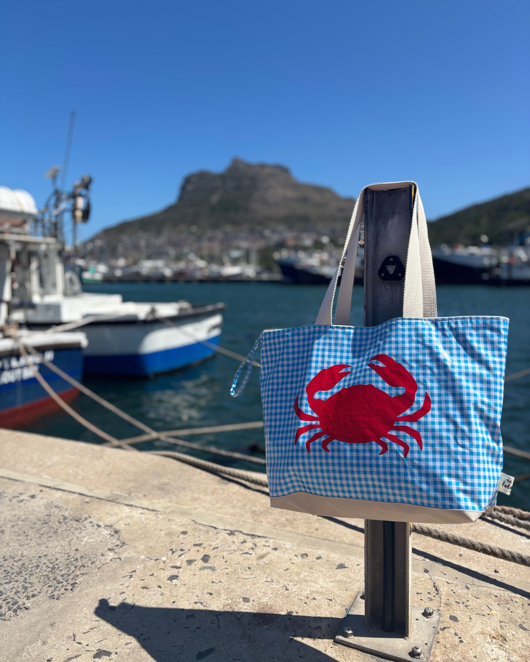 Blue gingham tote bag with red crab hanging on metal post at sunny marina with boats and mountain