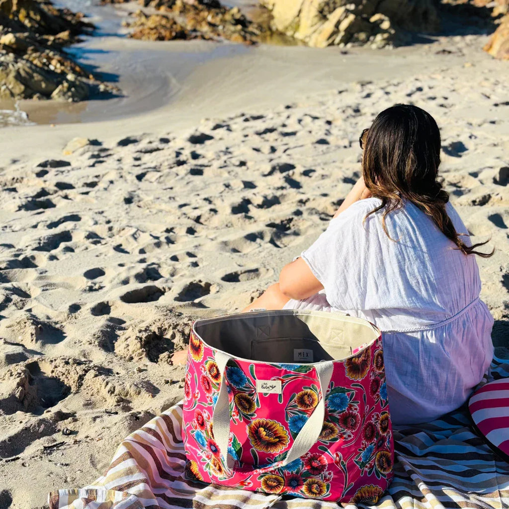 Woman in white dress sitting on striped beach towel with colorful floral tote bag on sandy rock beach