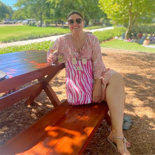 Woman in pink floral dress and sunglasses sitting on wooden picnic table bench, holding wine glass outdoors