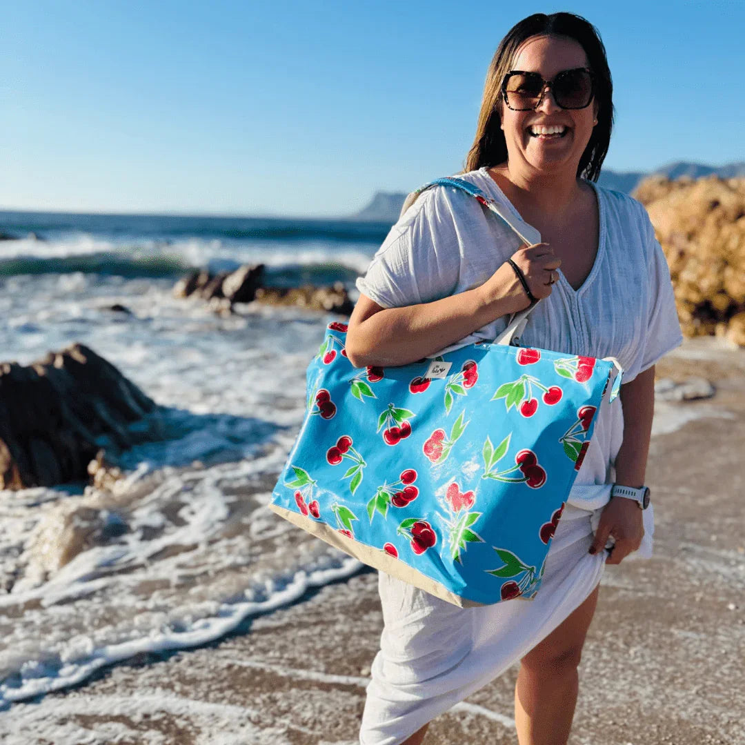 Smiling woman in white dress holding blue cherry print beach bag on the sandy shore with waves