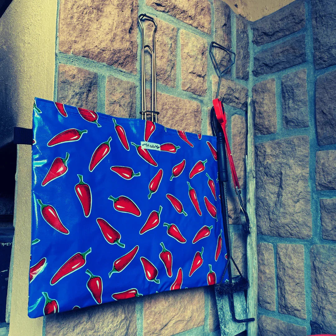 Blue apron with red chili pepper print hanging on stone wall with barbecue tools