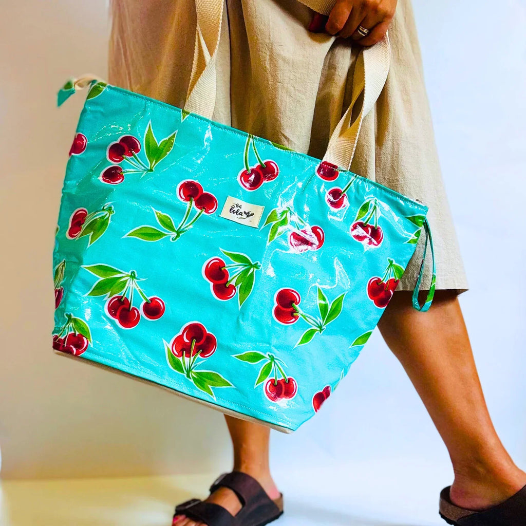 Person holding a turquoise tote bag with red cherry pattern and beige handles, wearing beige skirt and black sandals