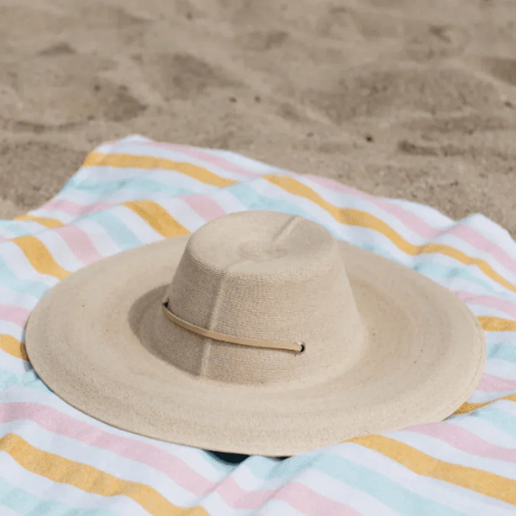 Beige wide-brim sun hat resting on striped pastel beach towel on sandy beach