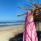 Pink and white striped beach towel hanging on dry thatched umbrella with sandy beach and ocean waves