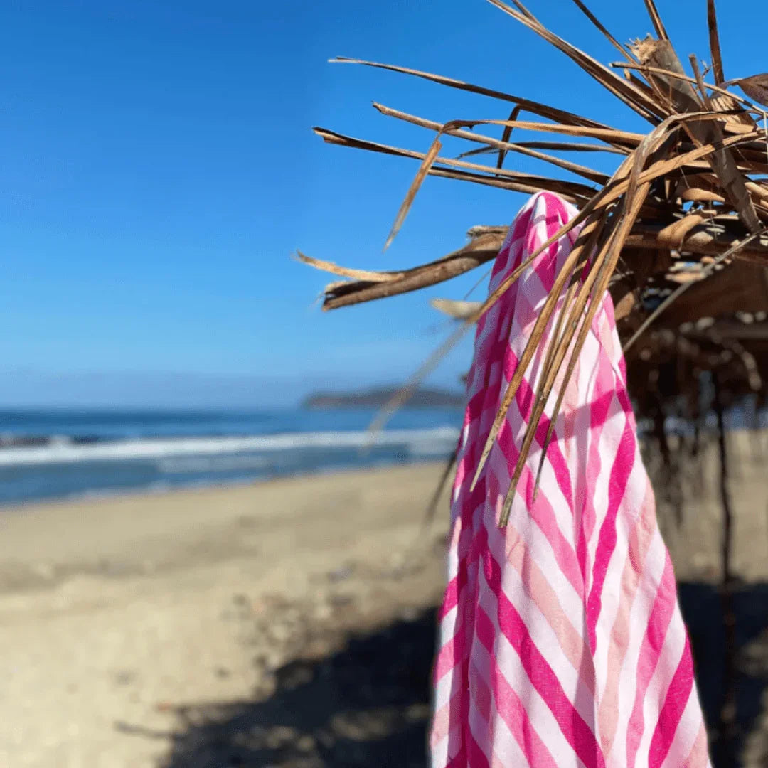 Pink and white striped beach towel hanging on dry thatched umbrella with sandy beach and ocean waves