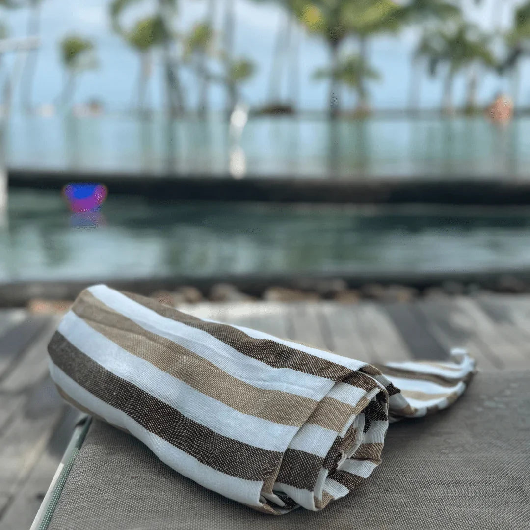 Rolled beige and white striped beach towel on lounge chair by pool with palm trees in background