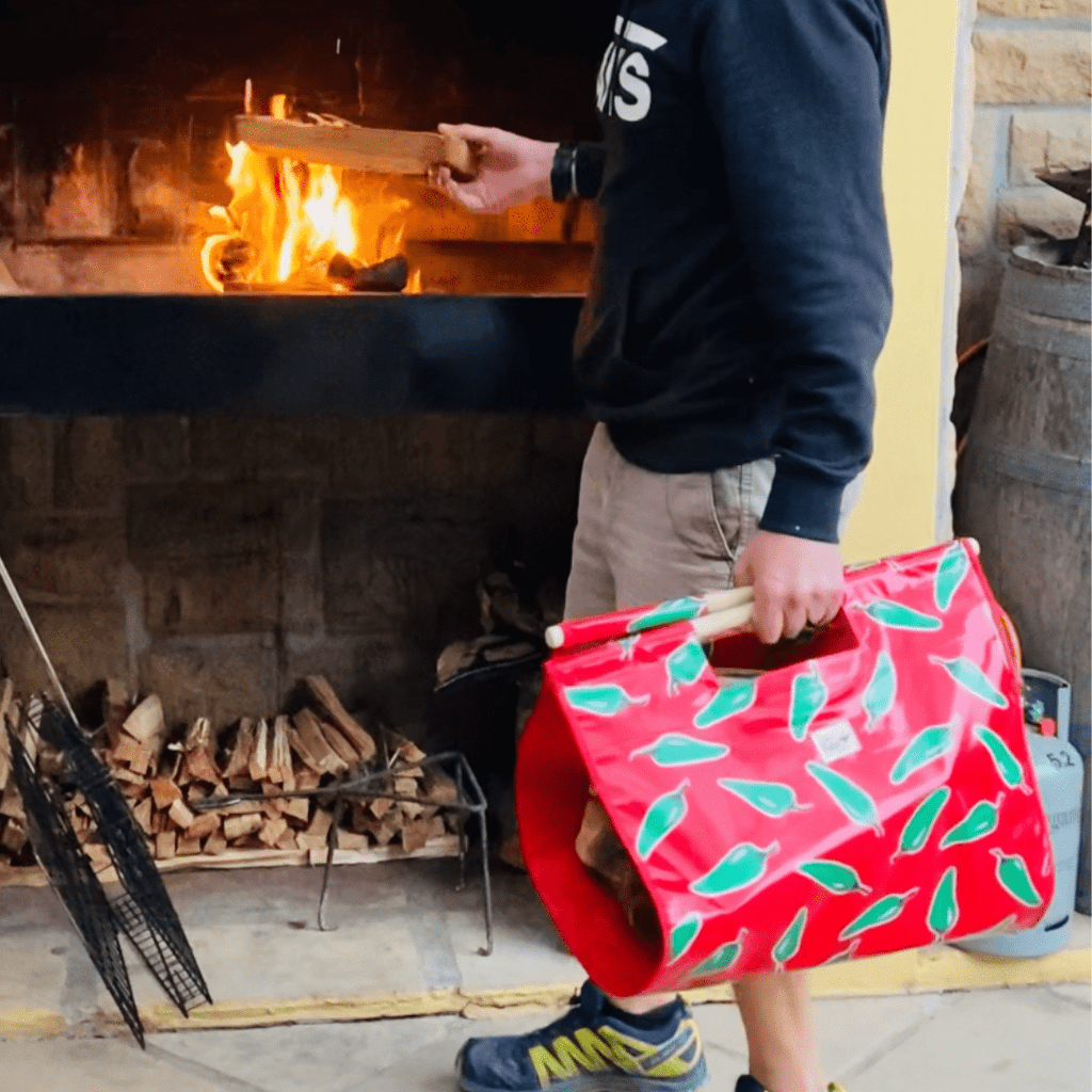 Person adding firewood to indoor fireplace holding a red firewood carrier with green leaves