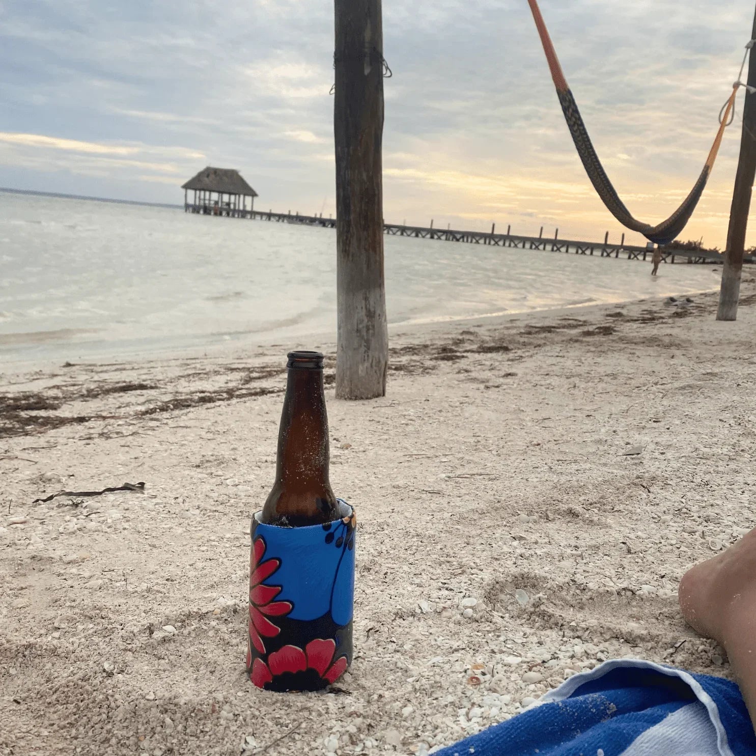 Brown beer bottle in blue floral koozie on sandy beach near wooden pier at sunset