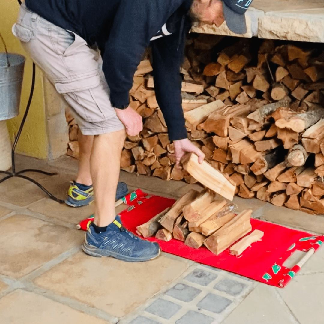 Person stacking firewood on a red mat near a stone fireplace with a woodpile
