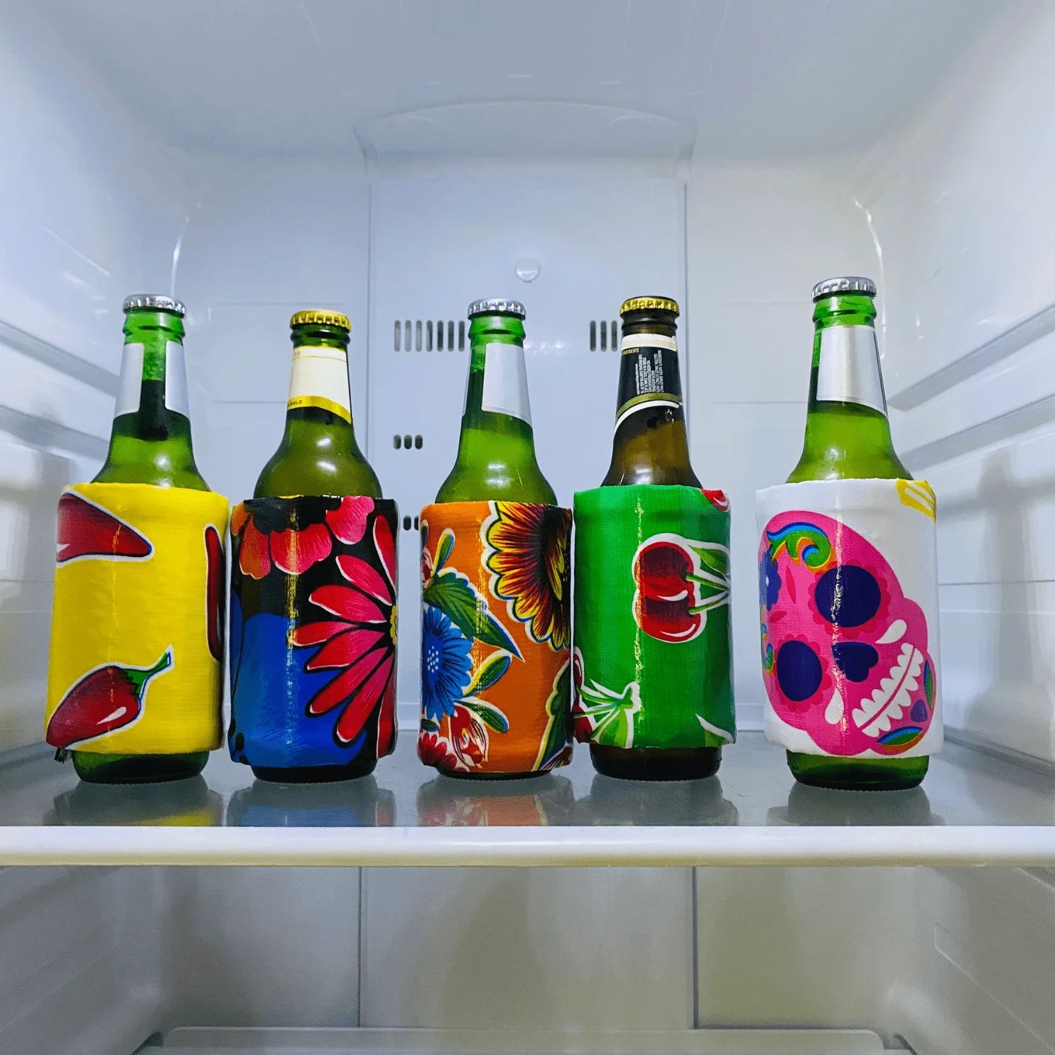 Five green beer bottles in colorful fabric koozies with chili, floral, cherry, and skull designs inside a refrigerator