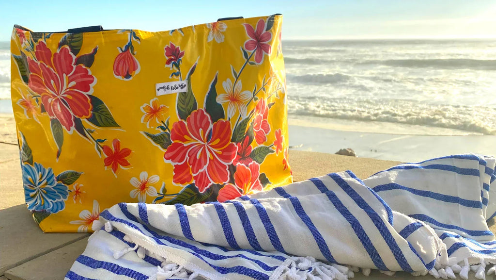 Yellow floral beach bag with red, pink, and blue flowers and striped blue towel on beach near ocean