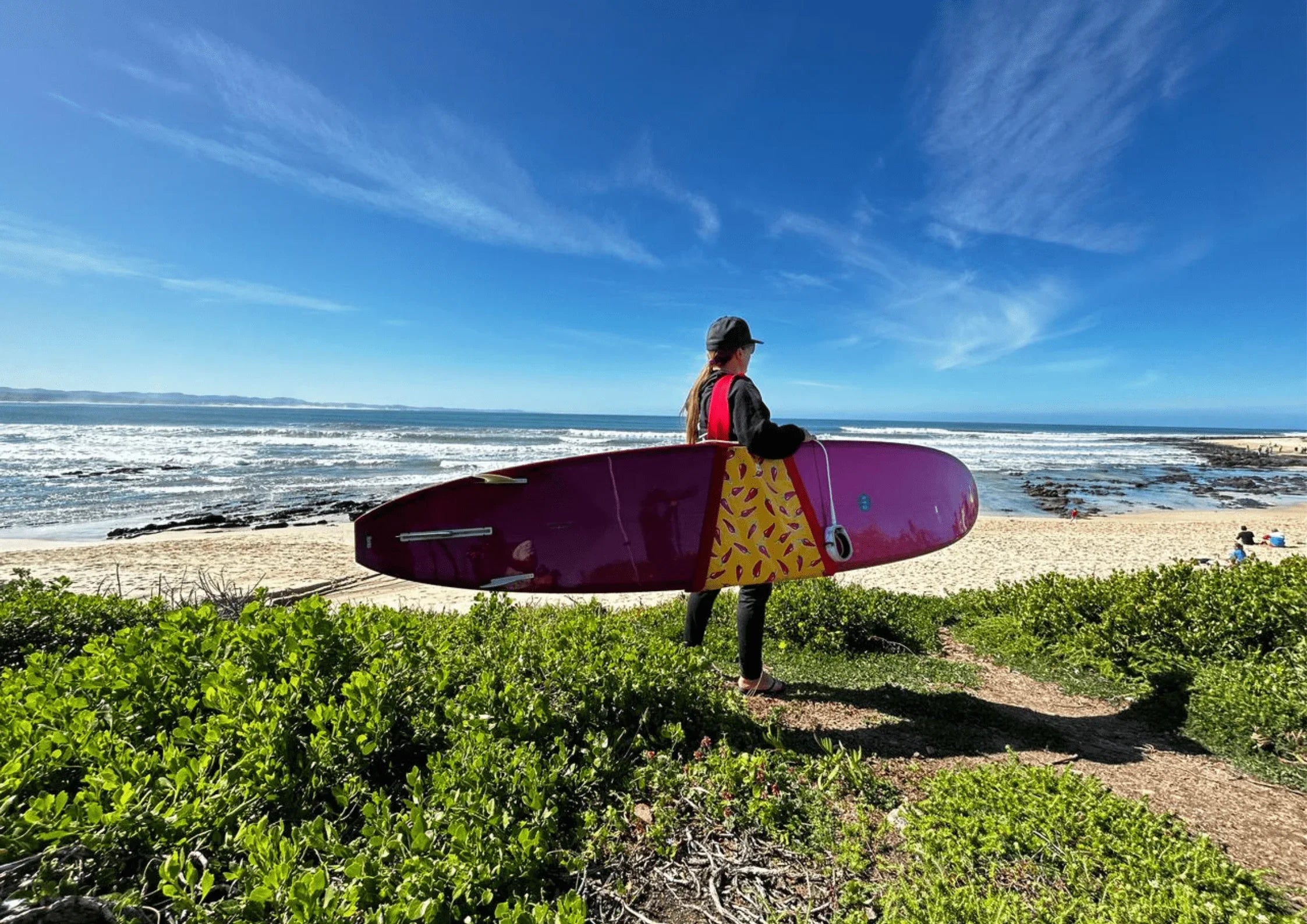 Person in black outfit and cap holding a purple surfboard with yellow design by beach under blue sky